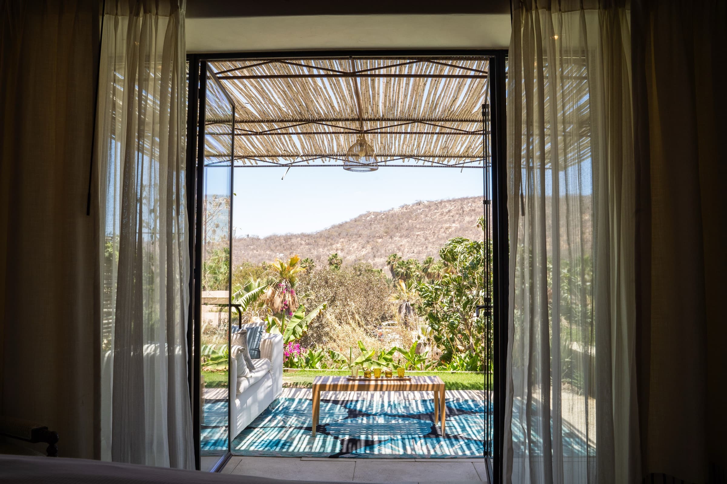 View from a Flora Farms hayloft bedroom through open steel-framed doors to a covered terrace with a striped rug, lounge chair, and Sierra de la Laguna mountain backdrop