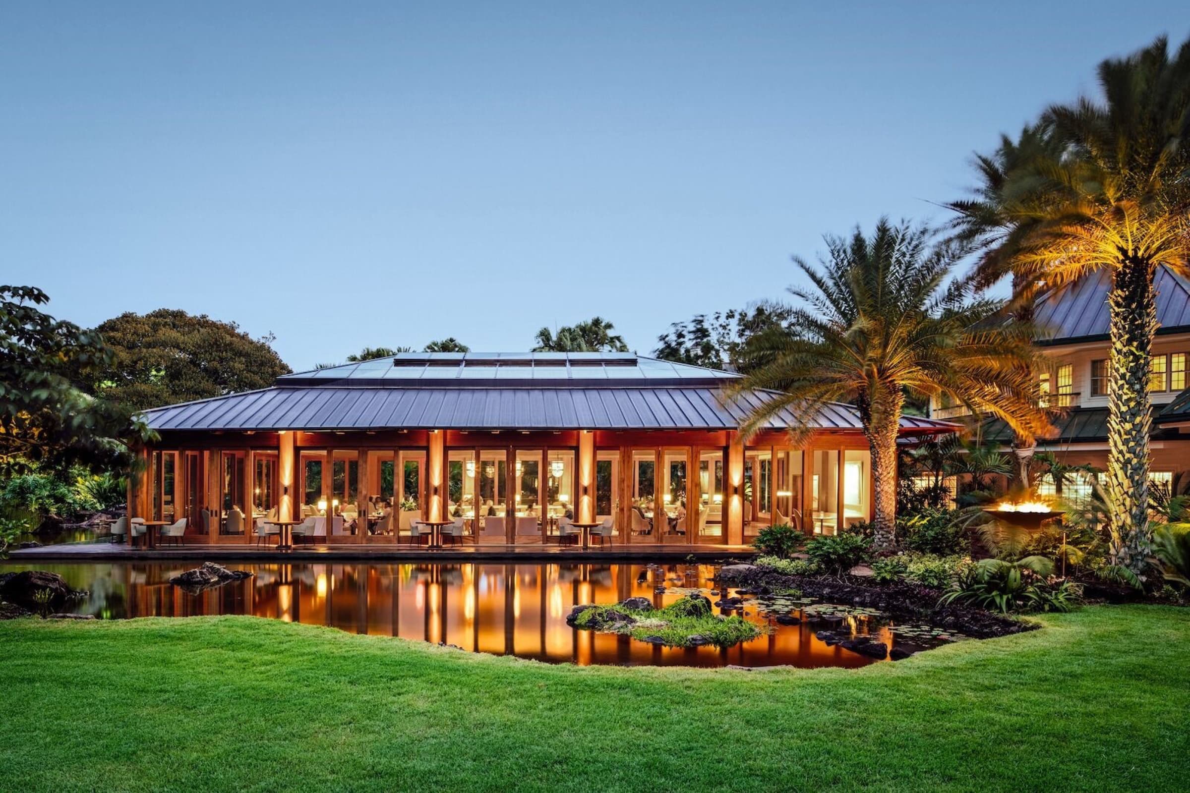 Resort restaurant building at blue hour: glass facade with warm interior glow, fan-shaped metal roof, reflected in a surrounding pond