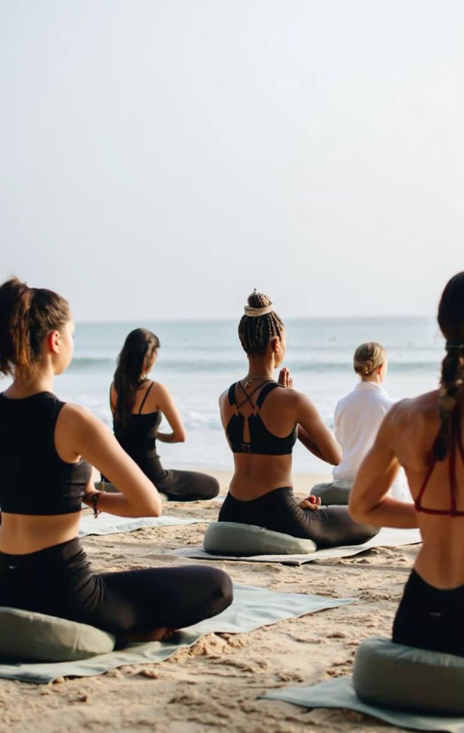 Women in a seated meditation on yoga mats facing the ocean at sunrise on the beach